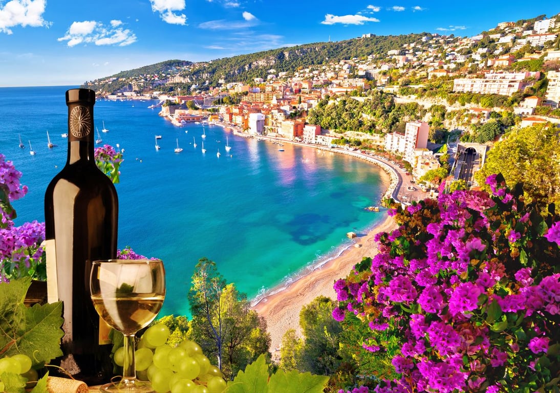 תמונה של בקבוק יין וכוס לצידם פרחים סגולים וברקע עיר וחוף ים