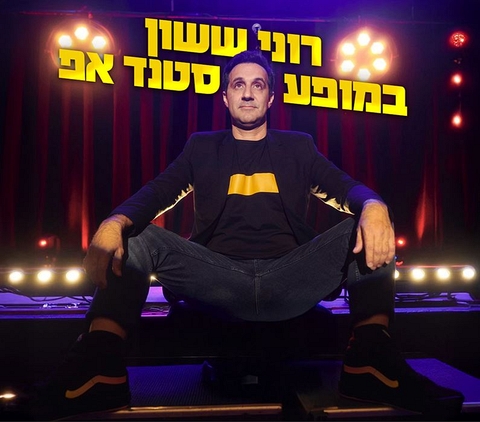 רוני ששון יושב על הבמה ברגליים פשוקות לצדדים