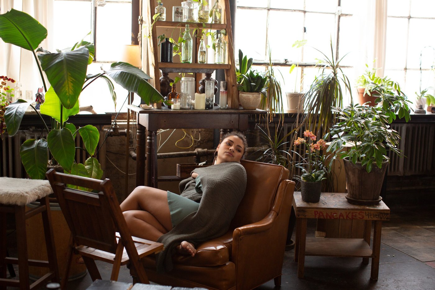 Thoughtful woman wrapped in scarf looking away while relaxing on armchair by potted plants at home