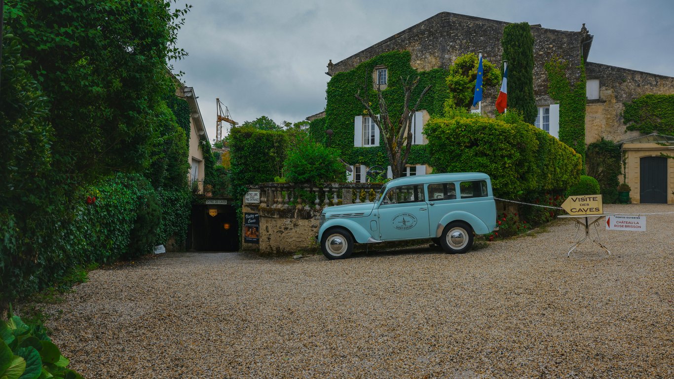 A romantic cobblestone square in Saint-Émilion, with historic stone buildings and a vintage car at its center, surrounded by the sounds of the town