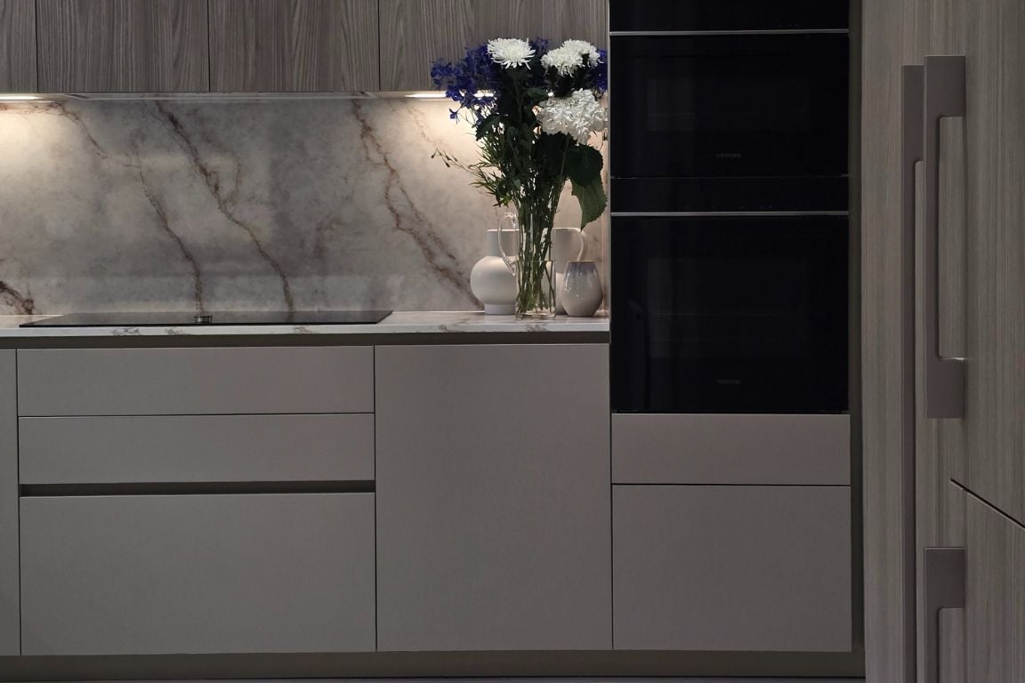 Close-up of a modern kitchen with marble backsplash, black ovens, and a vase of flowers.