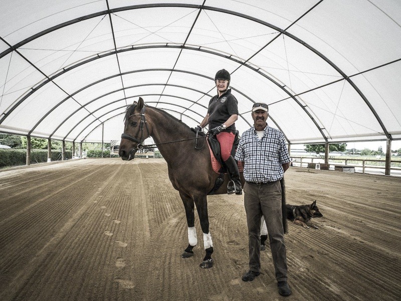 Indoor riding arena featuring a large brown horse with a rider seated atop, an older man standing beside them in a plaid shirt and cap, and a resting dog nearby on the groomed sandy ground, all framed by the curved white canopy of the structure.