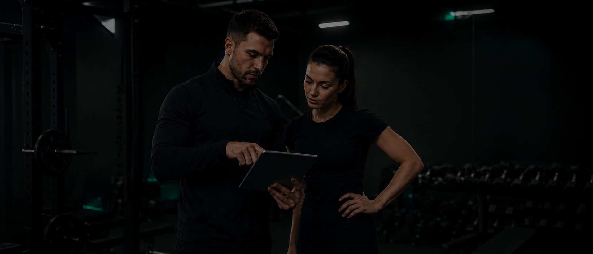 Treneris ir klientė sporto salėje peržiūri individualią sporto programą planšetėje