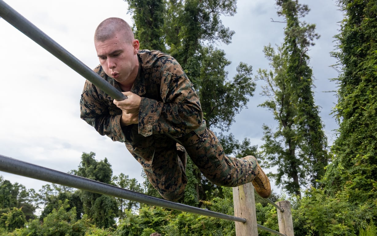 An NROTC Marine-option midshipman in Marine camouflage utilities crosses parallel bars on an outdoor obstacle course