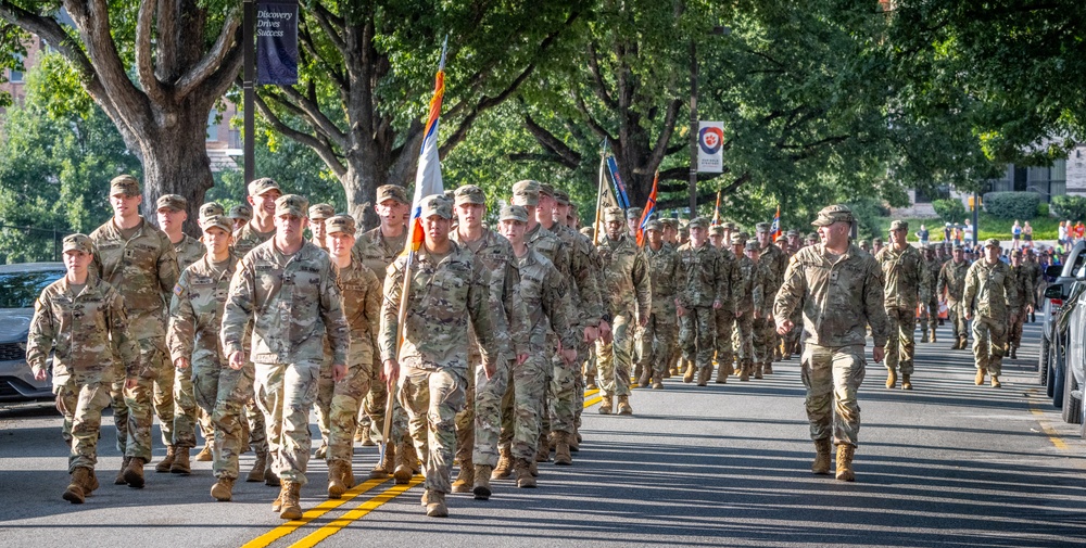 Army ROTC cadets in camouflage uniforms march in formation through a college campus, led by a color guard.