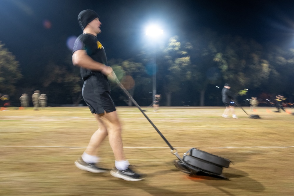 An Army ROTC cadet in physical training gear drags a weighted sled across a field at night under stadium lights during a scholarship fitness test event.
