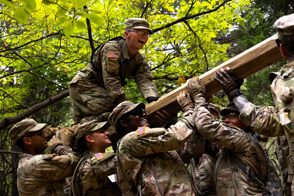 A team of Army ROTC cadets in camouflage uniforms work together to lift a heavy wooden beam over an obstacle in the woods on the Field Leader Reaction Course.
