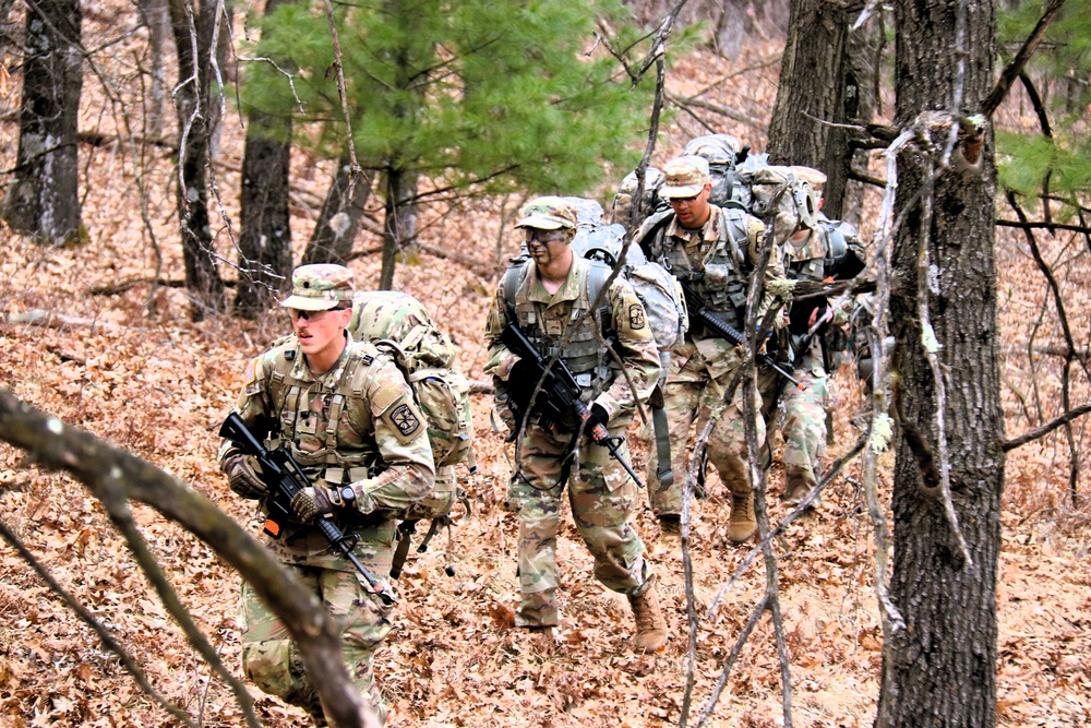 Army ROTC cadets from several universities in camouflage uniforms conduct small-unit field training in wooded terrain.