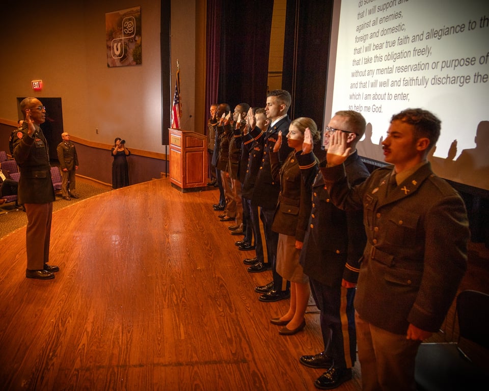 Army ROTC cadets take the oath of office during a commissioning ceremony, with the officer's oath displayed on screen behind them
