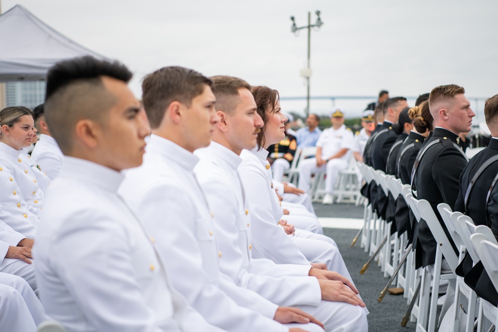 Newly commissioned officers in Navy dress white uniforms seated at a Navy ROTC commissioning ceremony.