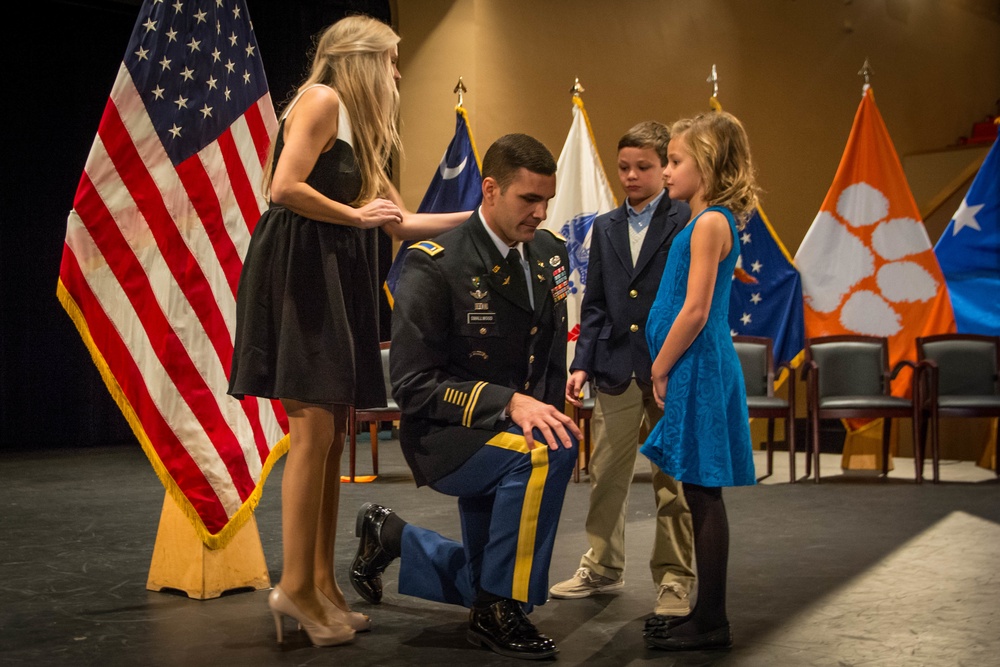 A new Army second lieutenant kneels as his wife and two children pin his gold bar rank onto his uniform at a Clemson University ROTC commissioning ceremony.