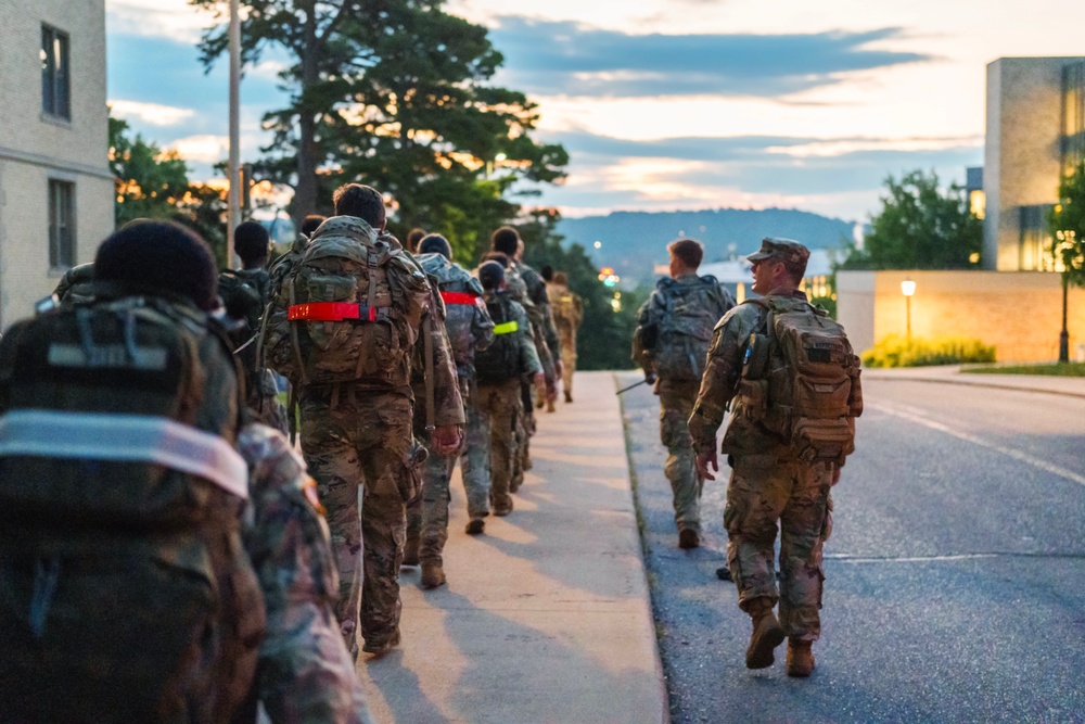 Army ROTC cadets in camouflage uniforms with rucksacks walk up a road at dawn during a ruck march, an instructor alongside them.