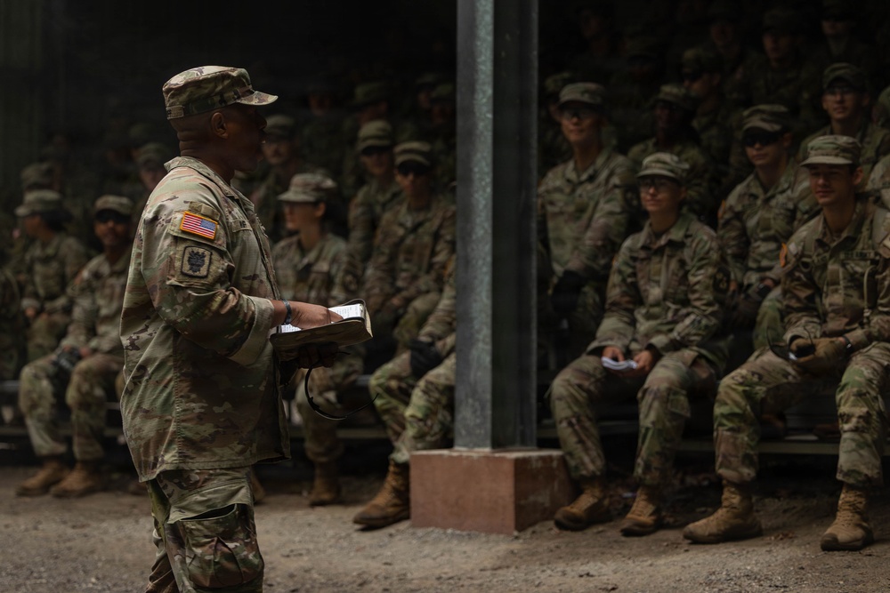 An Army cadre officer addresses a group of seated ROTC cadets in camouflage uniforms during summer training.