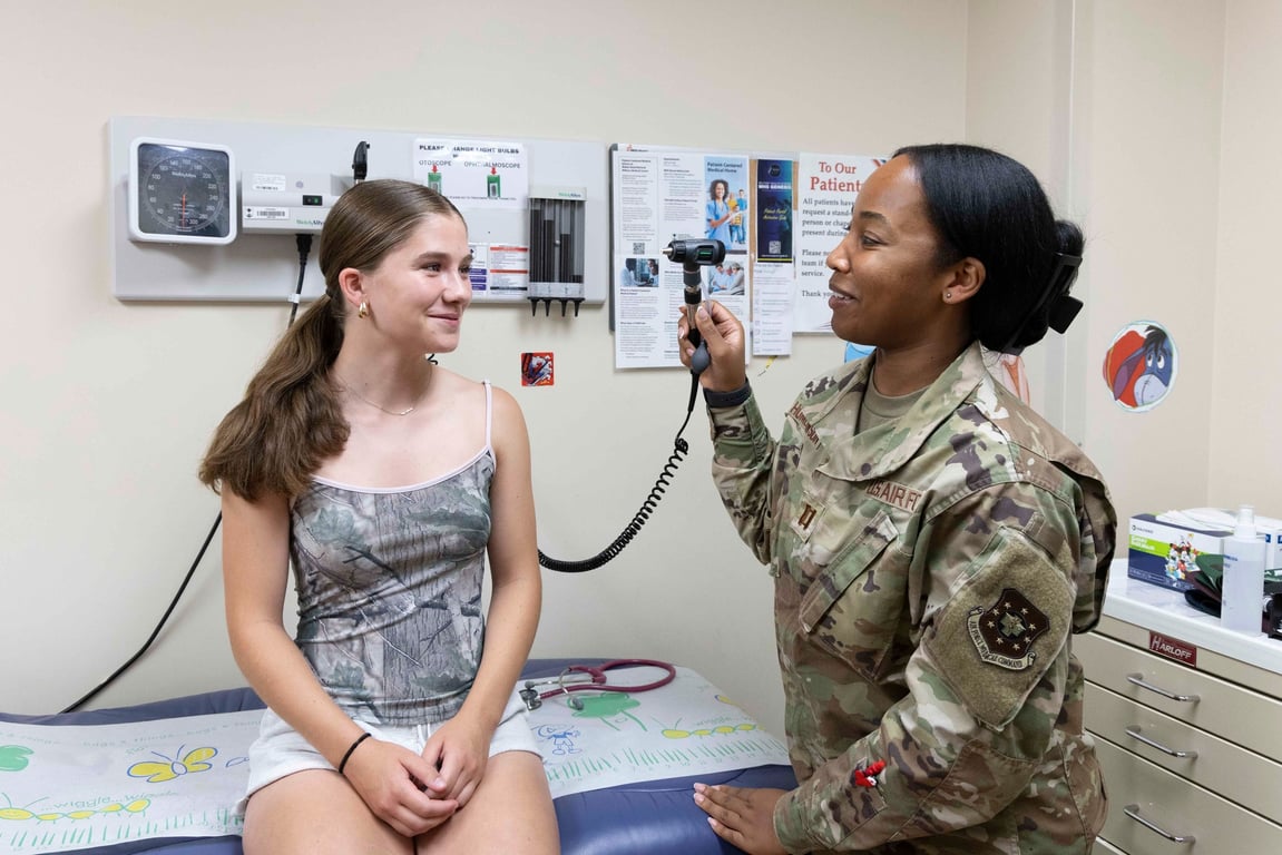 A U.S. Air Force medical officer in a camouflage uniform examines a young patient with an otoscope in a clinic exam room