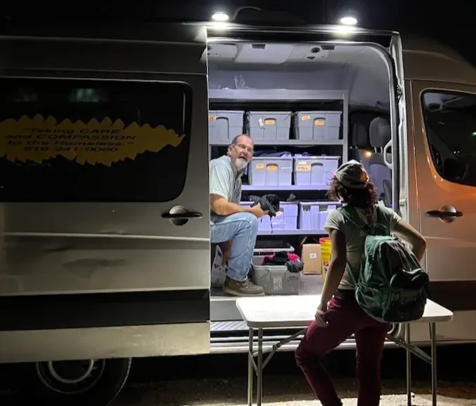 Volunteer kneeling beside an Uplift van at night, handing supplies to a person on the street. The van’s light shines into the dark, showing bins of food and clothing.
