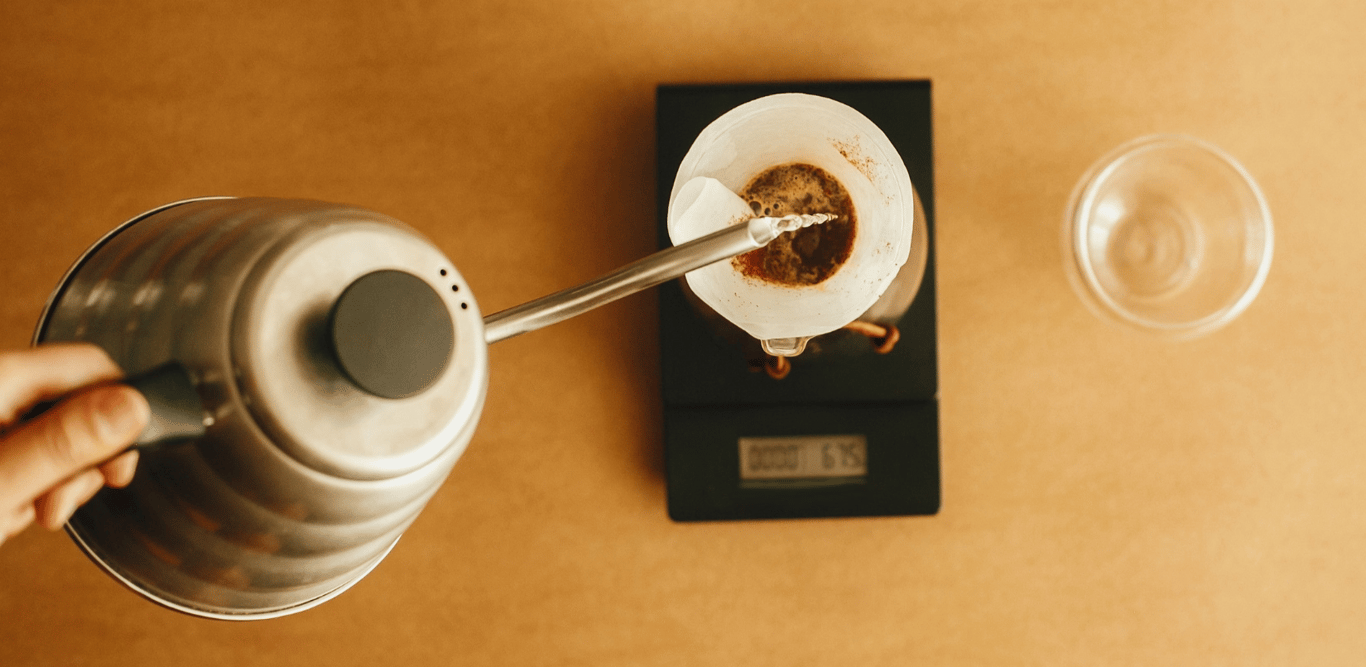 An overhead view of a hand pouring water into a pour-over filter resting on a scale, with a glass beside it — representing the precision and intentionality of strategic filtration