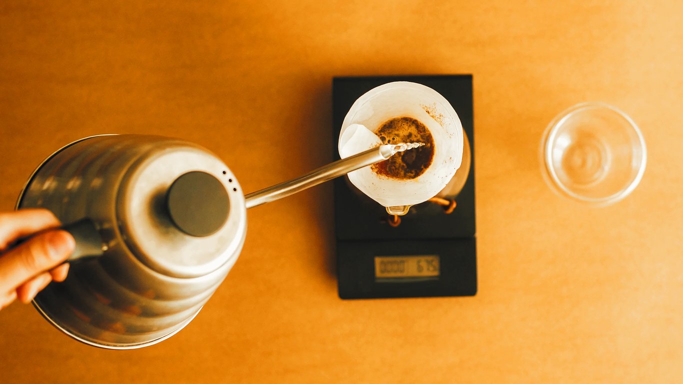 An overhead view of a hand pouring water into a pour-over filter resting on a scale, with a glass beside it — representing the precision and intentionality of strategic filtration