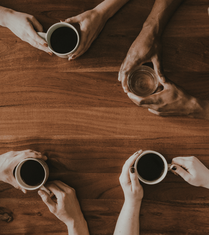 An overhead view of hands holding coffee cups around a wooden table, representing the collaborative, grounded spirit of The Common Table