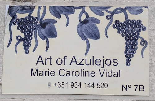White tile with blue vines of grapes and flowers that says Art of Azulejos, Marie Caroline Vidal. Includes phone number and address.