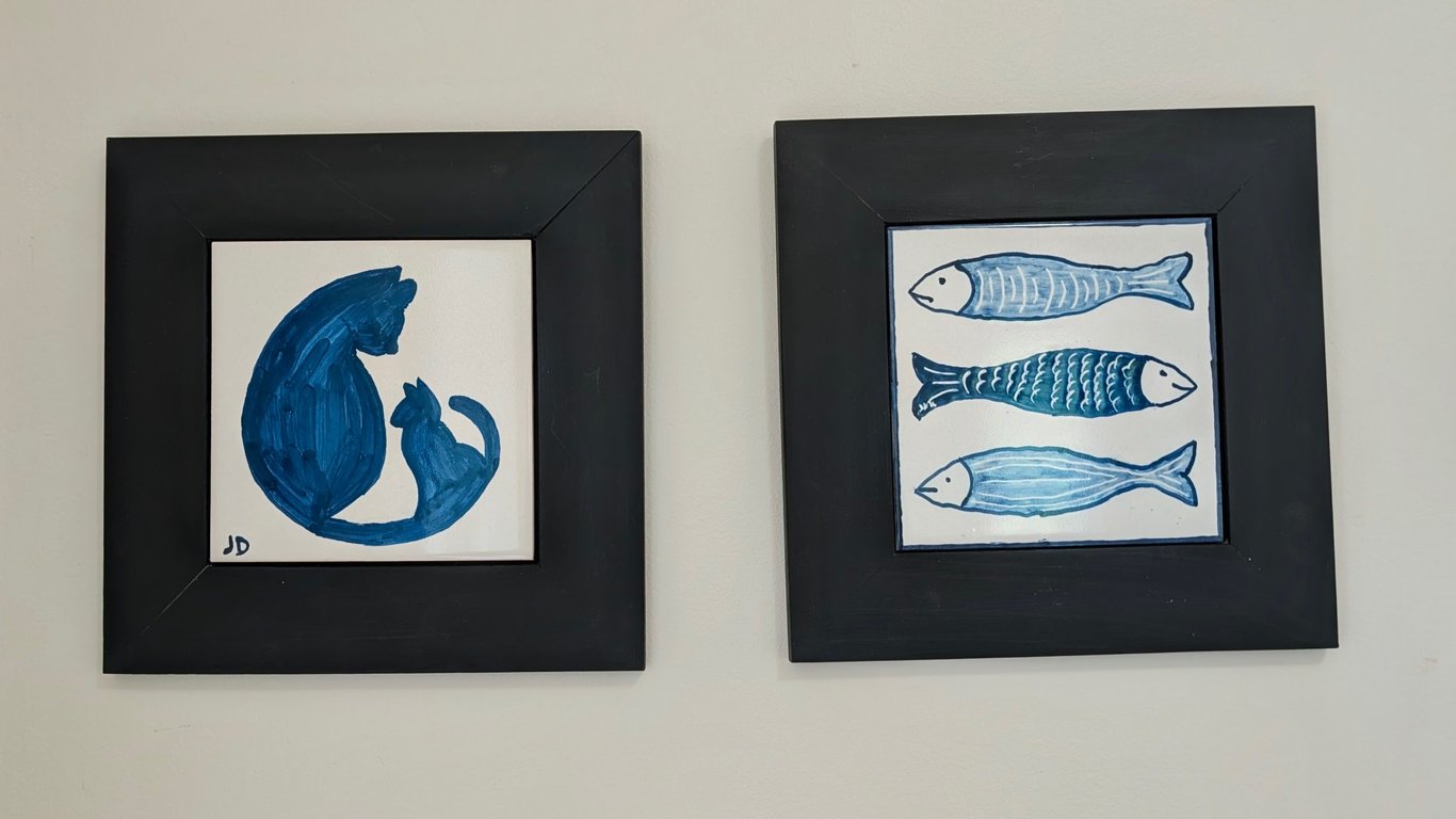 Two framed white tiles painted with traditional blue Portuguese paint. The tile on the left is of two cats looking at each other and the tile on the right is of three fish in a horizontal line.