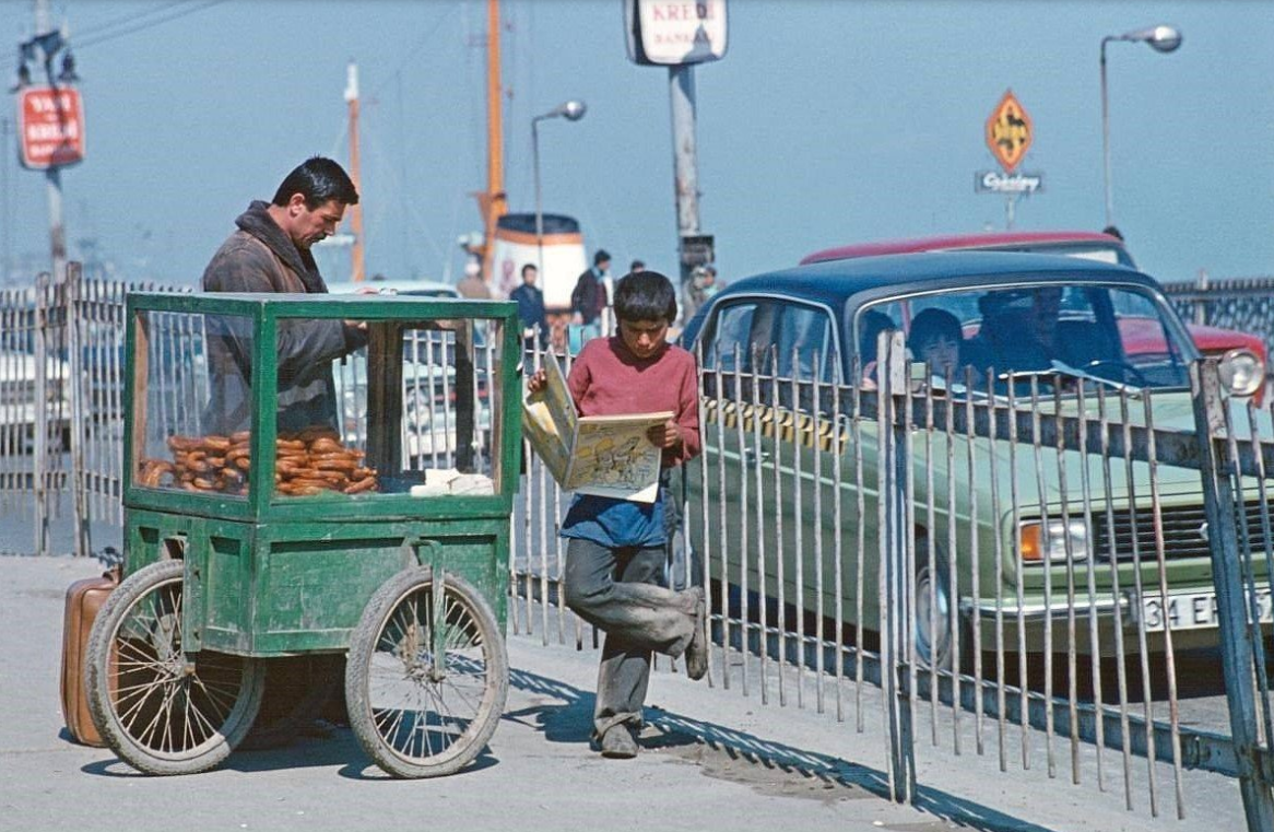    There are two man in the photo. And there are two people, one big and one small. I think he is a father and he one next to him is his son. The boy is holding a cartoon magazine in his hand. The man is a bagel seller. There are some bagels in the old green wooden bagel cart. And a suitcase is standing in front of the man. The suitcase is right at the bottom of the bagel cart. A three-wheel bagel cart. The boy rested one foot on the iron railing at the side of the road. There's a ferry in the back.