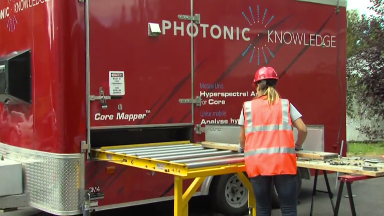 View of the outside of the mobile trailer imaging laboratory, the Core Mapper. A female engineer wearing a safety vest is loading a tray of rock core samples onto a roller conveyor which feeds to the inside of the trailer.
