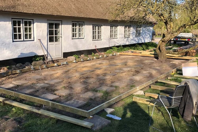 Foundation preparation for a wooden deck, showing a perimeter frame and paving stone supports laid out next to a thatched-roof house.