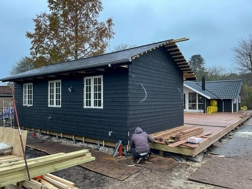 Person working on the construction of a dark wood-sided building elevated on piers, with an adjacent deck under construction.