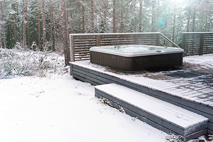 Winter scene featuring an outdoor hot tub on a snow-dusted deck, surrounded by snowy terrain and pine trees.