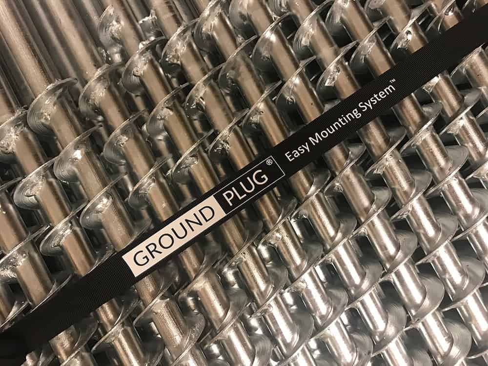 A close-up shot of multiple silver metal GroundPlug® screw piles arranged diagonally in rows. The piles have a helical flange design. A black strap with the text "GROUND PLUG® Easy Mounting System<sup>TM</sup>" is wrapped diagonally across the piles. The strap is centered in the frame, highlighting the brand name. The image suggests a product display or packaging of GroundPlug® foundation components.