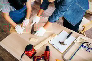 High-angle view of two individuals working together on carpentry; one marks wood using a square while the other assists, surrounded by tools.