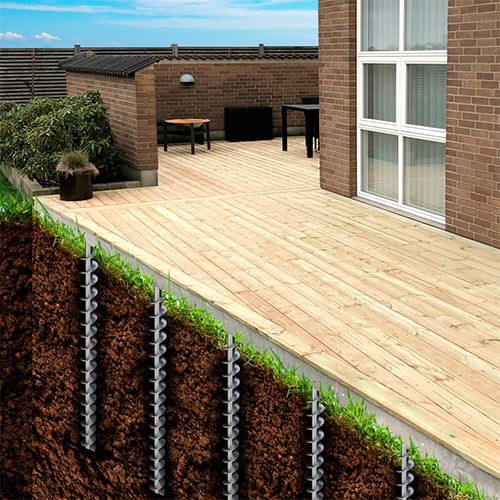 Cutaway diagram showing helical ground screws anchored in the earth, providing foundation support for an adjacent wooden patio deck connected to a brick building.