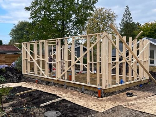 Framing stage of a building project, showing erected wood stud walls, floor system on piers, and initial roof structure assembly.