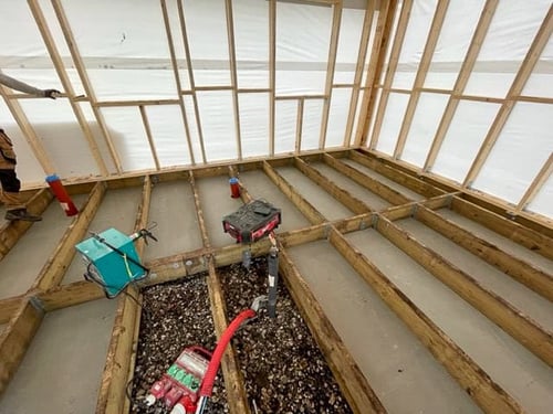 Floor system under construction inside a wood-framed building, showing bays between joists filled with gravel and smooth concrete, with red and grey plumbing pipes, red conduit, and power tools visible.