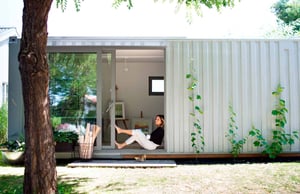 Person sitting in the open doorway of a modern, container-style building in a garden setting.