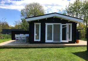 Modern black DIY garden room / cabin with white trim, large glass doors and windows, sitting on a deck with outdoor furniture.