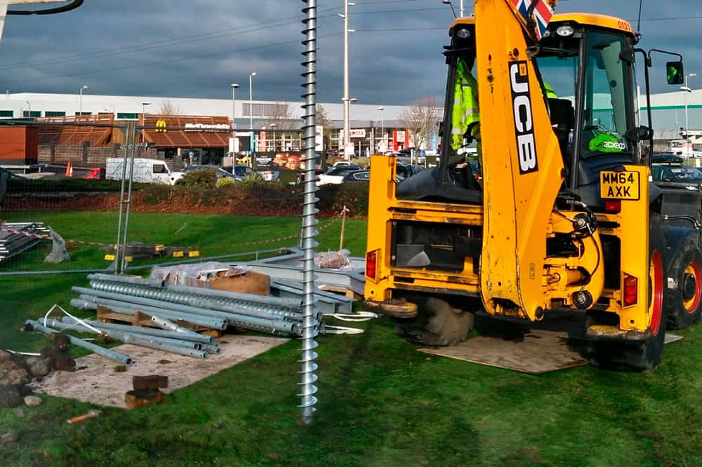 A bright yellow JCB 3CX backhoe loader is positioned on a green lawn, with a long GroundPlug® screw pile lying horizontally in the foreground. The screw pile is a silver metal auger, and a stack of metal poles is visible nearby. The JCB has "JCB" branding clearly visible, and "MX64 AXK" on the license plate. The background shows a commercial area with buildings and parked vehicles under a cloudy sky.
