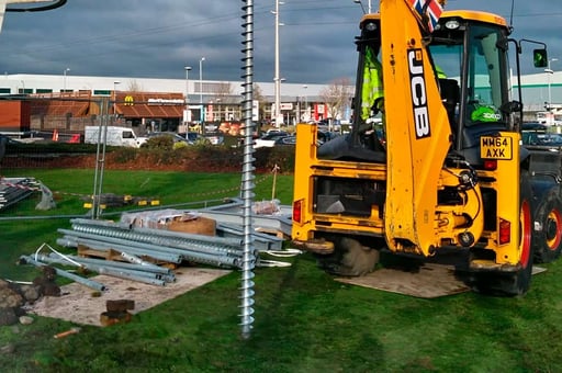 A bright yellow JCB 3CX backhoe loader is positioned on a green lawn, with a long GroundPlug® screw pile lying horizontally in the foreground. The screw pile is a silver metal auger, and a stack of metal poles is visible nearby. The JCB has "JCB" branding clearly visible, and "MX64 AXK" on the license plate. The background shows a commercial area with buildings and parked vehicles under a cloudy sky.