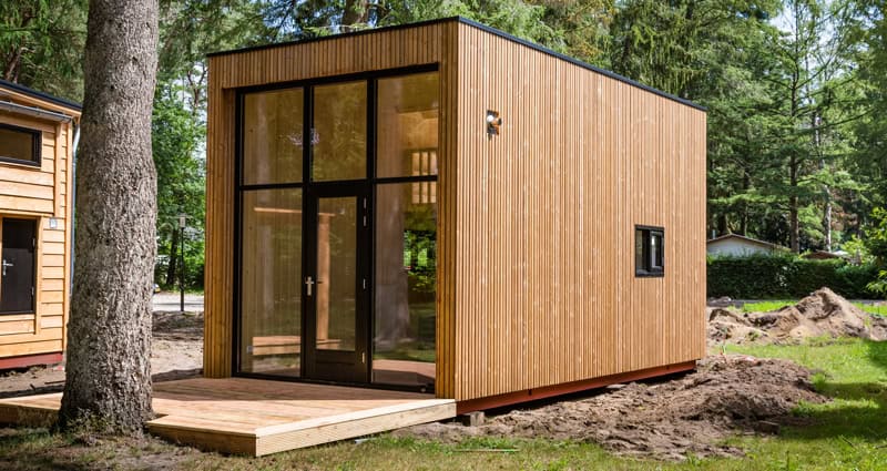 A contemporary, cube-shaped tiny house featuring vertical natural wood siding and a prominent black-framed glass entrance wall with a door, located in a clearing among trees.