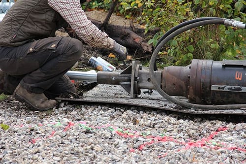 Construction worker preparing or connecting a heavy hydraulic drive head to a foundation element on a gravel surface.