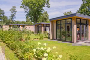 Multiple sleek, flat-roofed buildings with natural wood cladding and floor-to-ceiling glass windows, located in a well-maintained green space with plantings and trees. A year-round garden room.