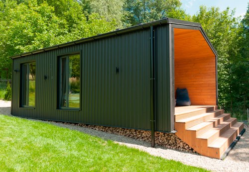 Modern tiny house or cabin design combining dark green metal cladding with warm wood accents on the recessed porch and steps, nestled into a green landscape.