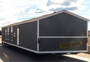 Section of a grey mobile or modular home being transported on a trailer on a highway, with a "BRED LAST" sign on the back.