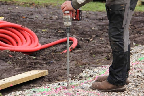 Installing a helical ground screw foundation using a handheld cordless power driver on a prepared gravel base, with orange conduit lying in the background.