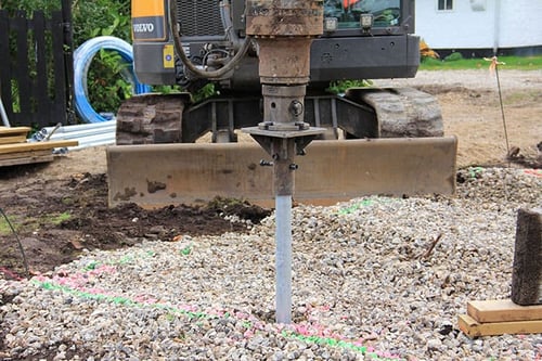 Excavator installing a ground screw foundation element into a gravel bed marked with paint.