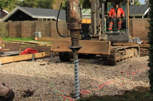 Excavator operated by a person in an orange jacket installs a ground screw on a gravel construction site.