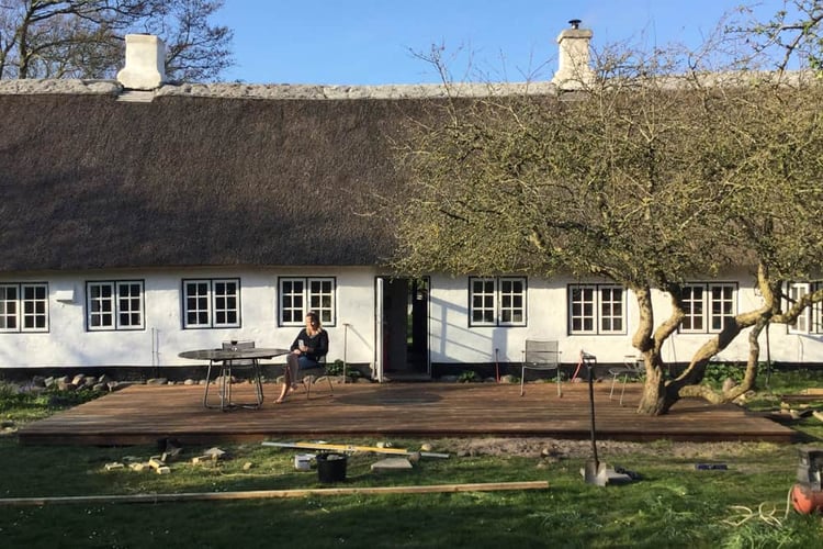 Person sitting on a wooden deck outside a long, white house with a thatched roof, with construction materials nearby.