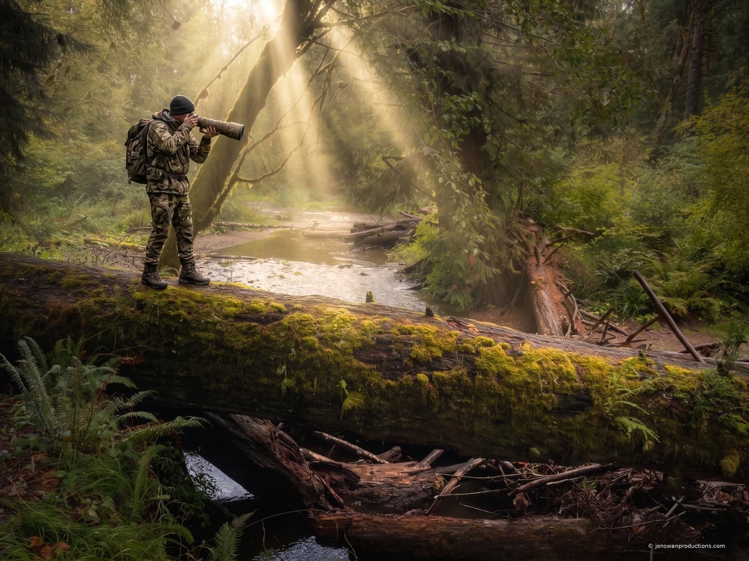 Photographer standing on a mossy fallen log over a creek, camera raised, sunbeams coming down through coastal conifer forest
