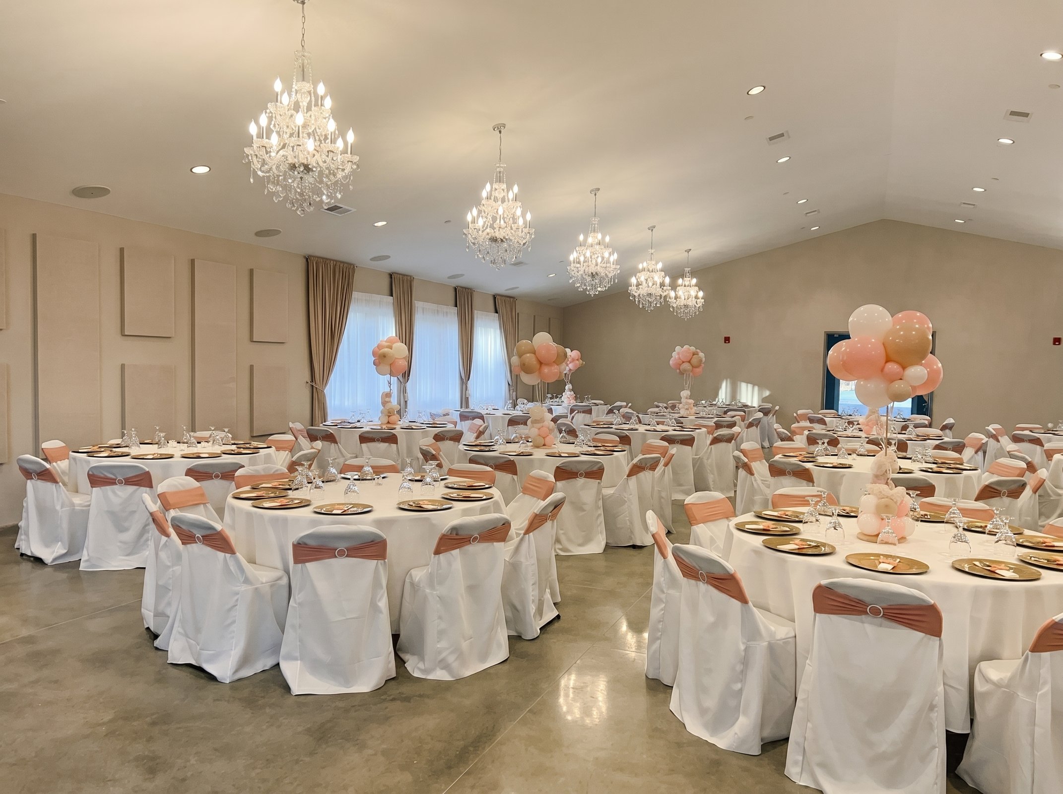 Round banquet tables with rose gold sashes, gold chargers and balloon centerpieces beneath the vaulted ceiling chandeliers