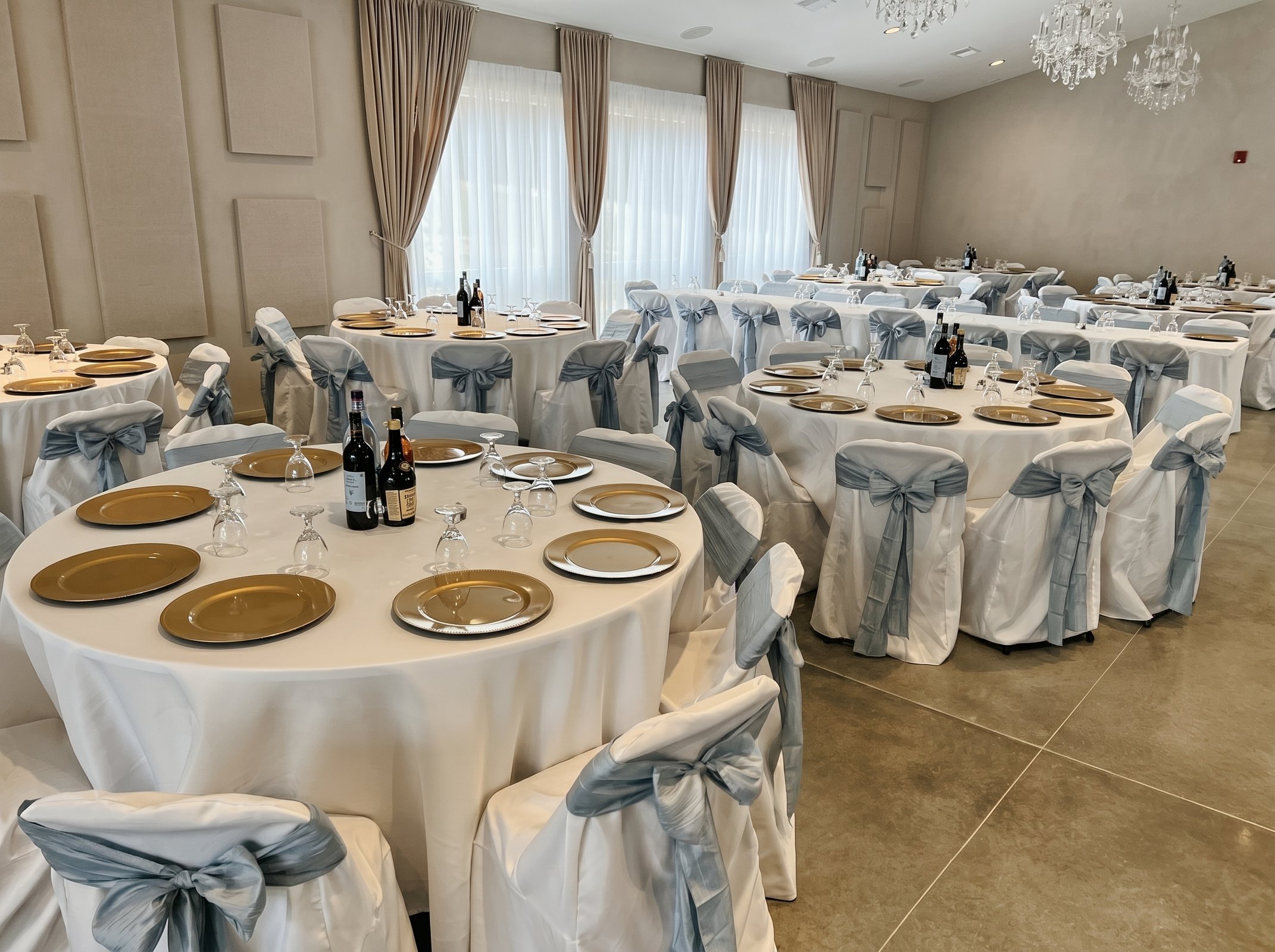 Wide view of round banquet tables with gold chargers and dusty blue chair sashes set for a reception